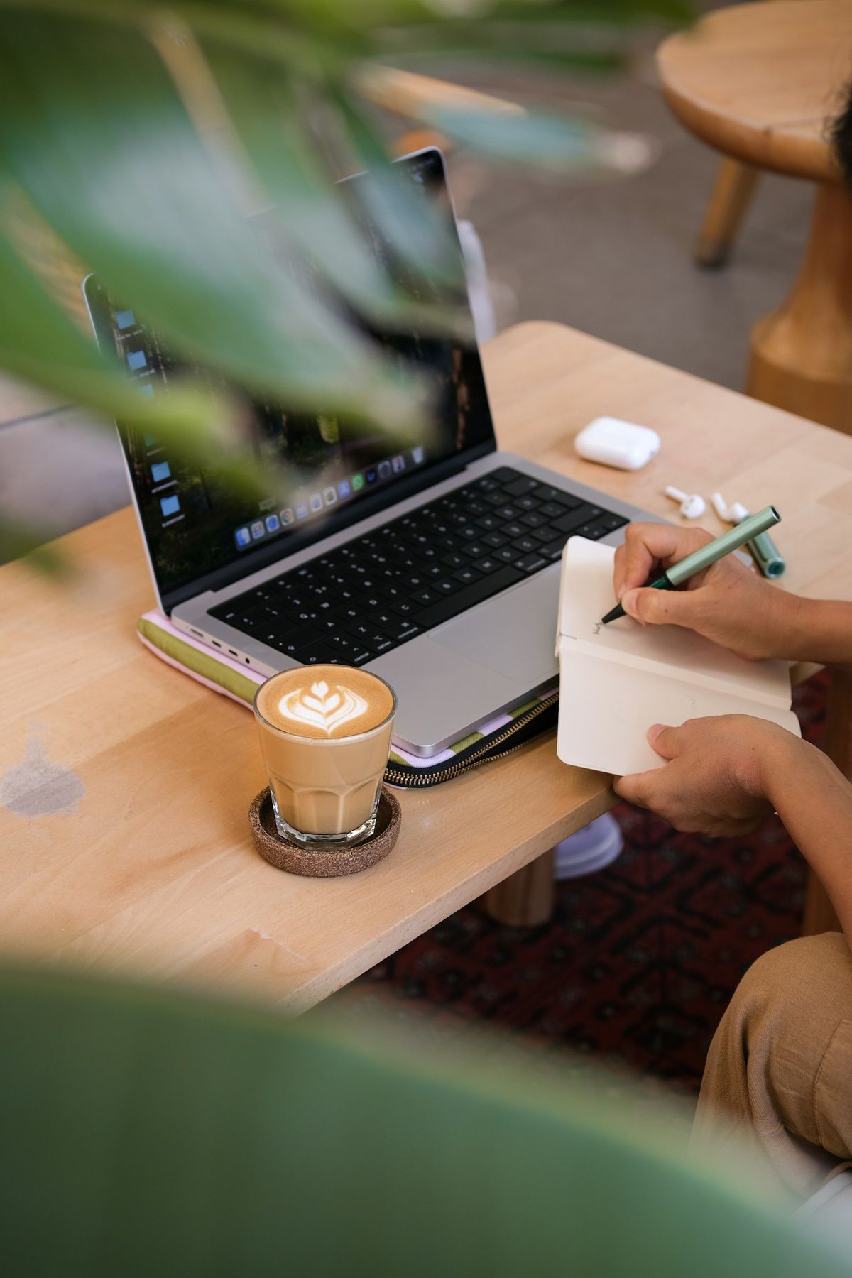 Person skriver i notatbok ved siden av laptop og kaffe